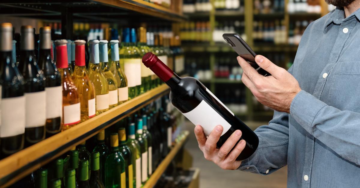 A man stands in the wine aisle at the liquor store as he holds a bottle of wine and scans the QR code label.