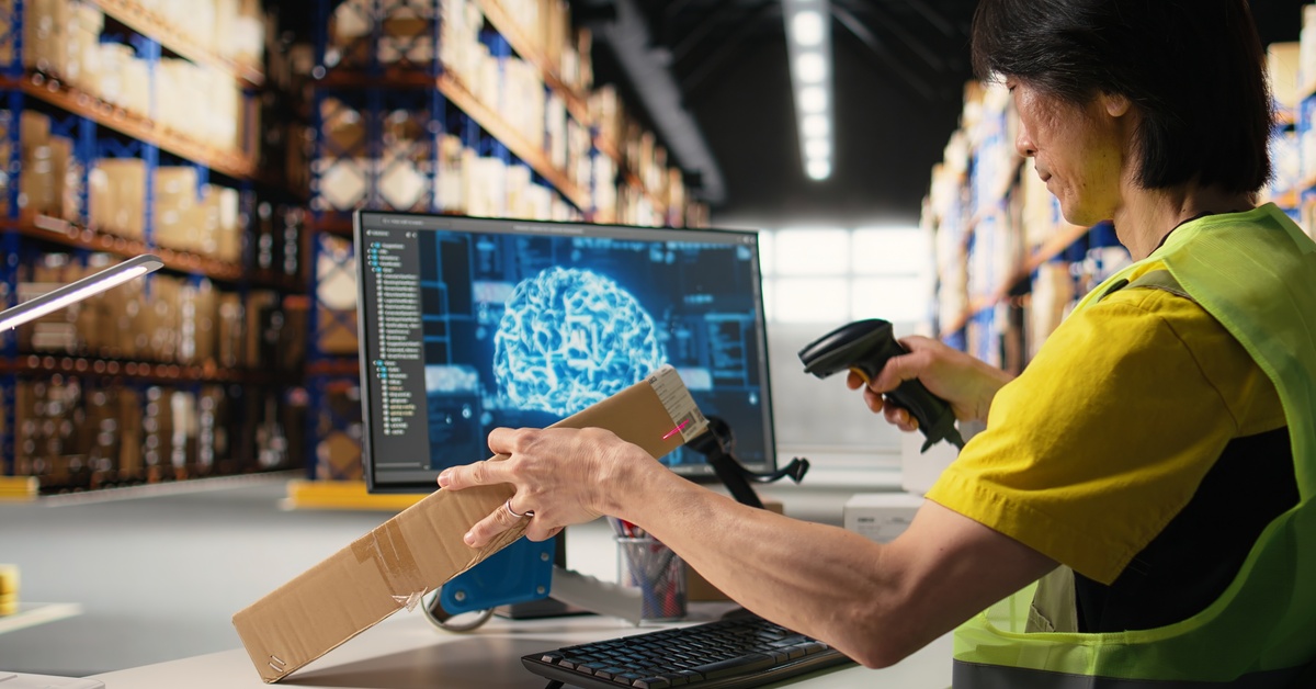 An aging warehouse employee sits at their desk scanning a box with the hand scanner. The computer screen is black with blue.