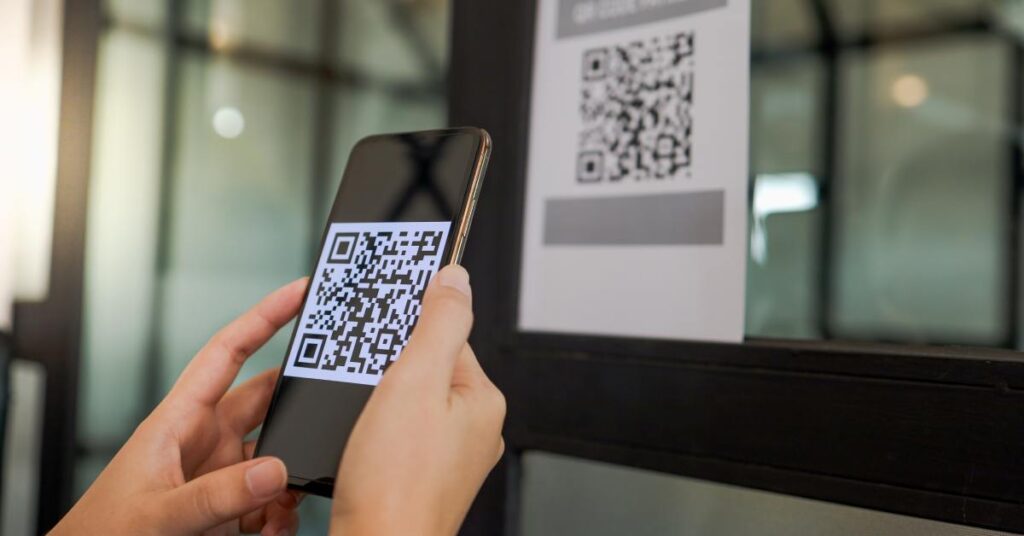 A person holds their phone with two hands near a white sign on the door that has a large black QR code.
