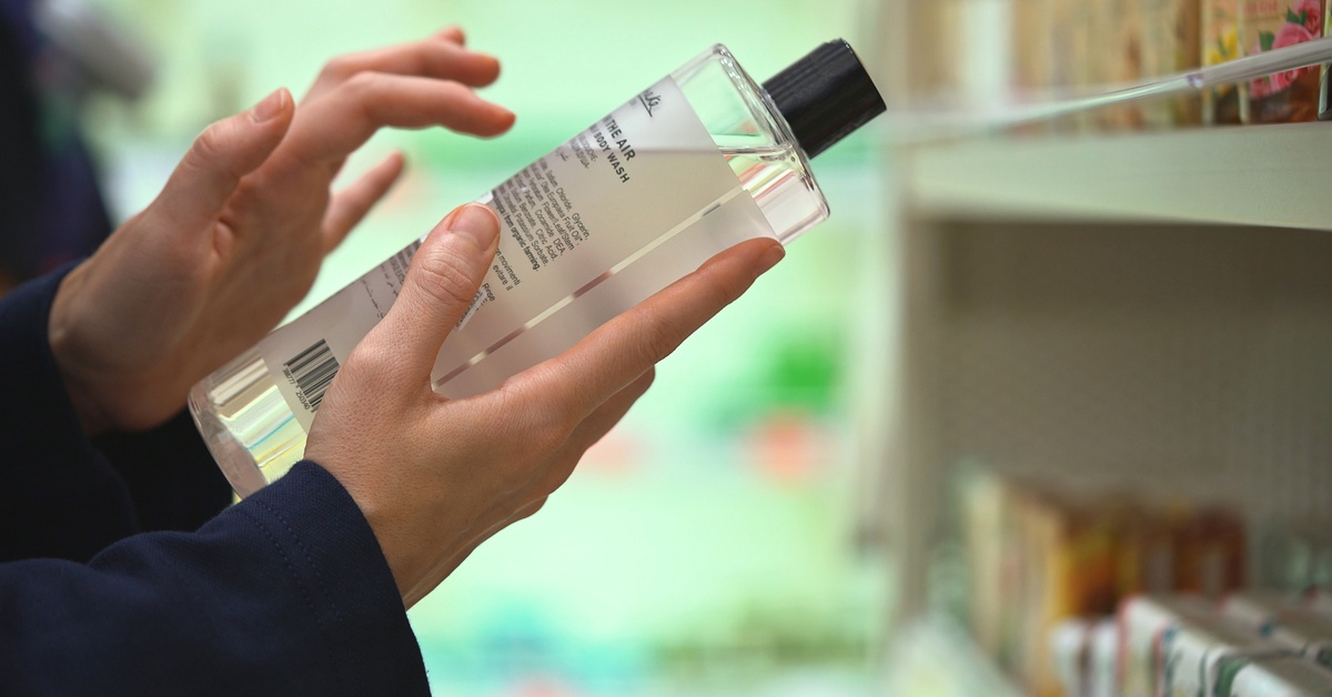 A consumer stands in the grocery store aisle and holds a clear bottle of soap. They scan the label with their fingers.