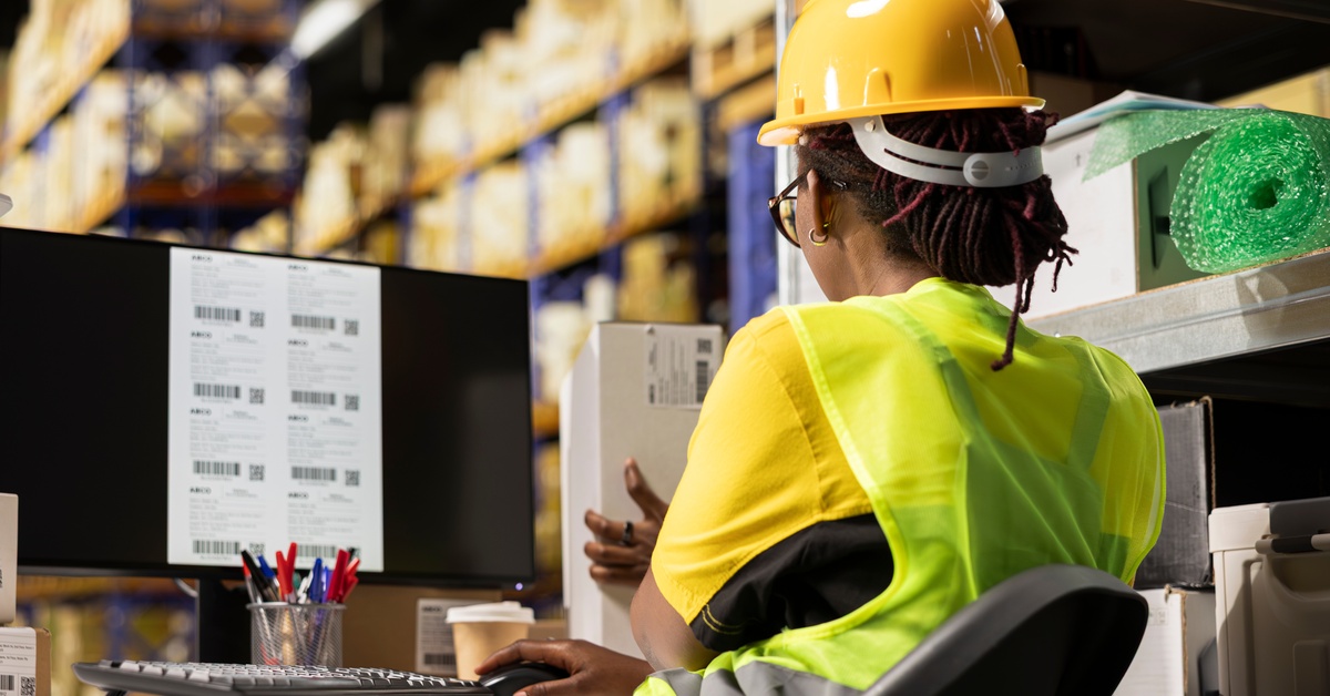 An employee in a yellow vest and hard hat sits at a desk making labels for boxes. The labels have barcodes.