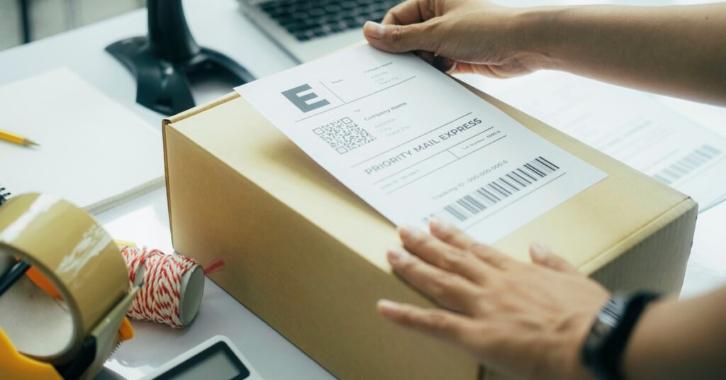 A close-up of a warehouse employee's hands placing a label on a small box. The label says "priority mail express."